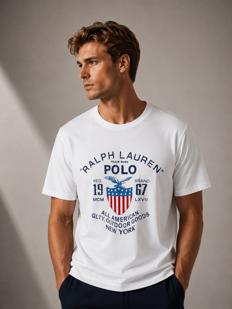 A man wearing a white short-sleeve t-shirt with a vintage-style Ralph Lauren Polo graphic on the front, paired with dark navy pants, set against a neutral gray studio background.