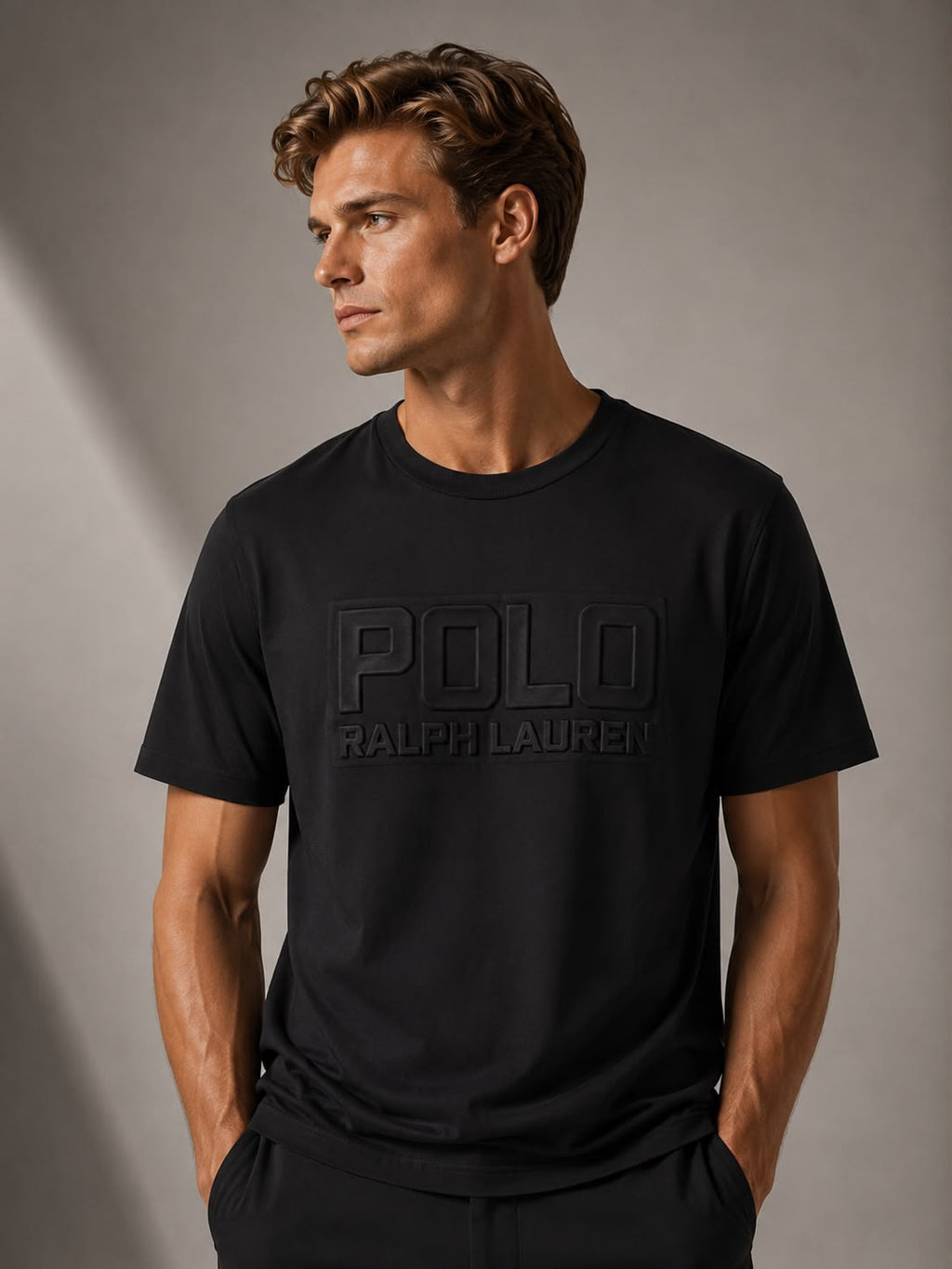A man wearing a black Polo Ralph Lauren t-shirt with a tonal black logo on the chest, standing against a gray studio background.