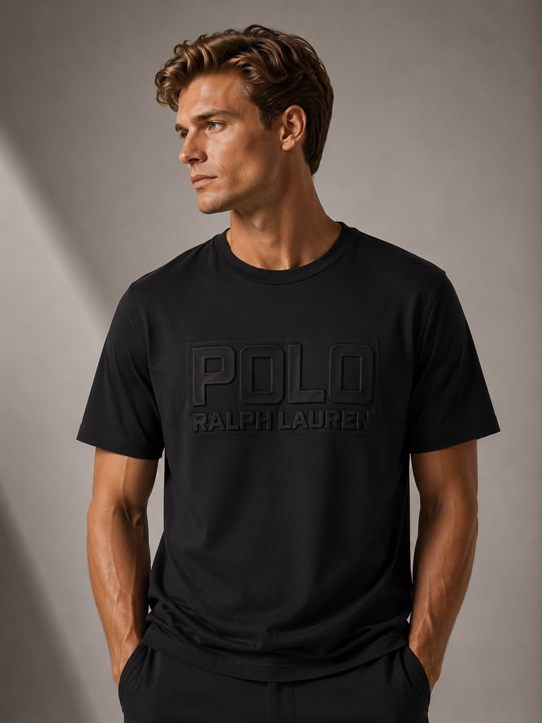 A man wearing a black Polo Ralph Lauren t-shirt with a tonal black logo on the chest, standing against a gray studio background.